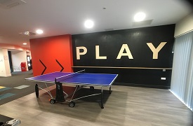 A room with a ping pong table in the centre and the word 'play' written on the wall.