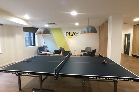A room with a table tennis table and two small coffee tables with several armchairs around them. The word 'play' is painted on one wall.