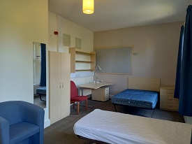 A single bed to the right in the foreground. Another single bed in the background. Wardrobe and desk in the far left corner of the room.