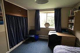 A room with a desk and office chair in one corner and a bed opposite it. There is a set of shelves over the desk and a cupboard covered by curtains on the opposite wall.