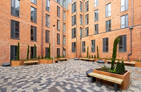A paved courtyard with benches and plant beds, enclosed on two sides by a large five-storey red brick building.