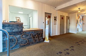 A hallway with a reception desk, hand sanitizer stand, and several doors leading off. The reception desk has 'DSW' written on it.