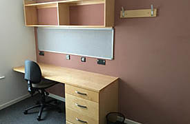 A desk in the corner of a room, with an office chair next to it and a set of shelves on the wall above it.
