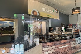 A counter with food in glass displays and a till behind a clear plastic screen. A sign on the wall behind the counter says 'Welcome to source Langford'.