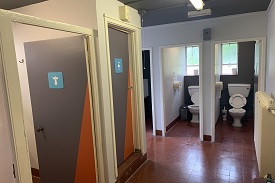 A room with two toilet cubicles, a door with an image of a shower head on it, and another door with an image of a basin on it.