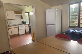 A room with a fridge freezer, recycling bins and a table with several chairs around it. There is a doorway leading into a kitchen which has another fridge freezer, an oven and hob, and several cupboards and drawers.