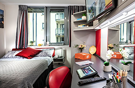 A room with a double bed in one corner and a desk and chair opposite. The desk has a mirror and shelves on the wall above it, with a television screen attached to the shelves.