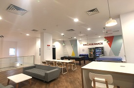 A large room with two sofas around a coffee table, a larger table with six chairs around it, a ping pong table, a pool table, and two vending machines against the far wall. The words 'fancy a snack' are written on the wall above the vending machines.