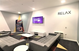A room with two sofas and an armchair around a coffee table. There is a television on the wall and the word 'relax' written on the wall nearby.