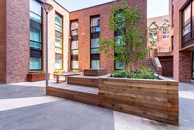 A paved courtyard with a raised wooden platform and seating, a ping pong table, and several plants in wooden holders. The courtyard is enclosed by a three-storey red brick building, with steps with a handrail leading up to a gate out.