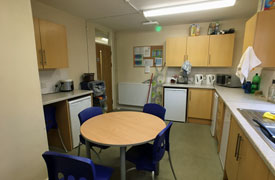 Shared kitchen area showing sink, several refrigerators, two kettles, toaster, microwave, storage cupboards, recycling bins and a table with four chairs