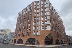 View of the residential block from street level. The brick building has 9 stories and a large archway entrance with double doors.