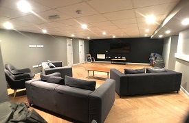 Cinema room with three sofas, two armchairs, a coffee table, recycling bins and a large screen at the far end of the room