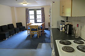 Communal kitchen and eating area showing cooker and hob, kettle, microwave, and storage units. At the far end of the room are a table and six chairs, and four armchairs