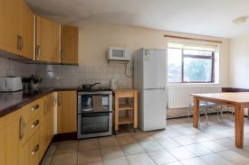 A kitchen/diner with a full-height fridge/freezer, an oven, kettle, toaster, and cupboard storage. There is a dining table by the window with stools placed alongside.