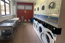 A laundry room with four washing machines and four tumble dryers alongside a wall. On the opposite wall with there is a double sink and countertop. On the far wall there is an electric iron and ironing board.