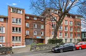 Street view of Woodland Court from Belgrave Road. The building is 5 stories high, and is positioned behind a brick wall and tall trees.