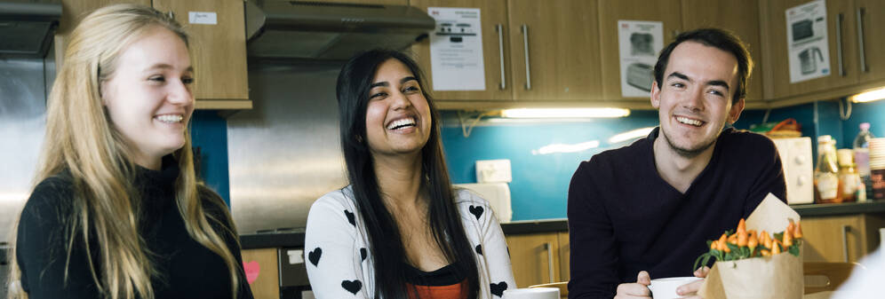 Three students sitting at a table laughing with each other in a kitchen in a self-catered student accommodation. 