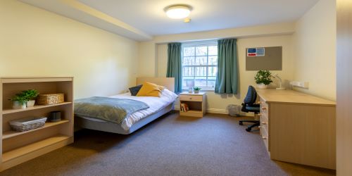 A student room in Churchill Hall, with a single bed in one corner and a desk opposite. A green space outside is visible through the window.