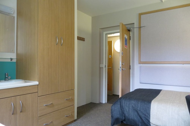 A room with a bed against one wall and a wardrobe, basing and cupboards against an adjacent wall at the foot of the bed.
