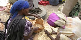 Two African women crouched down. One has a paper bag open while the other is passing rice/grain over to the paper bag.
