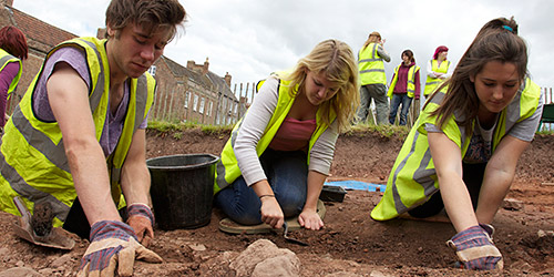 Three students knelt down, trowelling in the earth at an undergraduate excavation site. Several students are stood in the background.