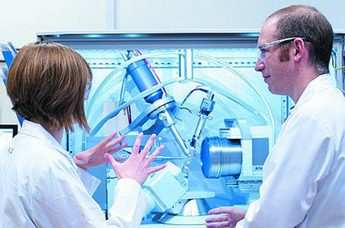 A man and a woman in white lab coats and goggles, looking at some complex machinery.