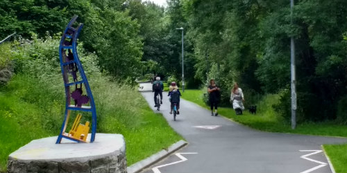 Two cyclists and two pedestrians travel on the Bristol to Bath cycle path. The path is surrounded by grass and trees and there is an abstract sculpture in the foreground.