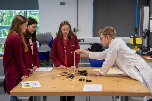 A group of three Sixth Form students are standing at a lab table with a CDT student, who is holding an aerofoil to their wing design, giving advice on their wing.
