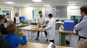students in white lab coats giving a demonstration to schoolchildren in a classroom