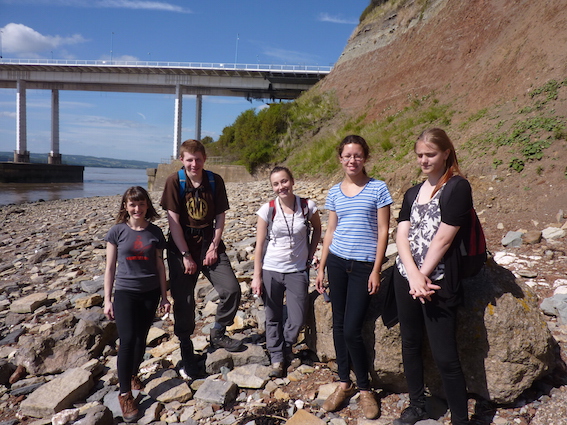 A group of five student interns standing on rocky terrain, enjoying a sunny day of field work on the Triassic-Jurassic boundary at Aust Cliff, near Bristol.