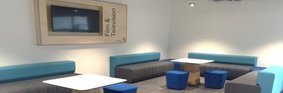 Four sofas with turquoise backs and grey seats with three small blue round stools gathered around white square tables portraying the 5th floor foyer area of Department of Film & TV