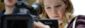 A female student looking into/focussing a camera lens.  A male student, wearing headphones and holding a long-armed microphone beside her.
