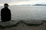 View from behind an older man sitting alone on a coastal wall, looking out over a tranquil grey sea.