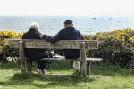 Elderly couple sitting on a bench in the countryside looking out to sea on a bright day. 