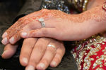 A close up image of a woman's henna-covered hand resting on her husband's hand on his knee. The couple are wearing wedding rings and are dressed up formally. She is wearing a beautifully embroidered red garment.