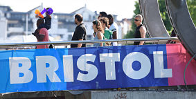 A group of people of different genders and races crossing Pero's Bridge in Bristol Harbourside. The word 'Bristol' is written across the bridge.