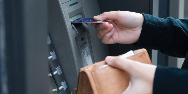 A person using a cashpoint; one hand is holding their wallet, the other is putting a card into the machine.