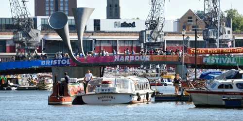 Boats passing each other under Pero's Bridge in Bristol Harbourside, with the MShed is visible in the background. The bridge has 'Bristol Harbour Festival' written on it.