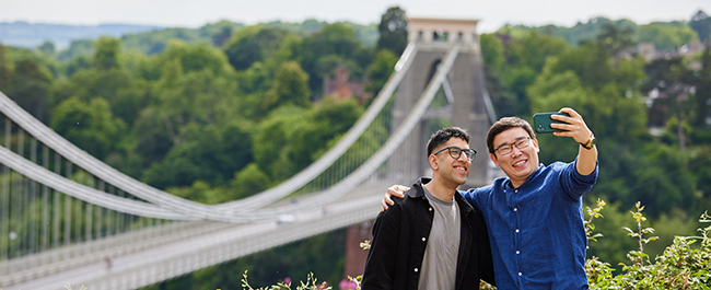 Two male students talking a selfie in front of the Clifton Suspension Bridge.