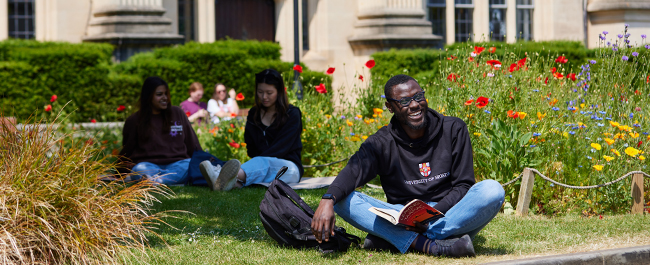 Male student sitting on the grass with a book, looking up and smiling. Bright flowers and other students sitting in the background.