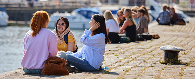 Three female students sitting on the harbourside chatting and laughing in the sun.