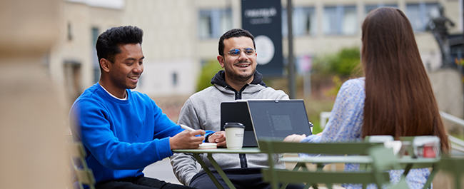 Three students sitting outside at a café working on laptops and smiling.