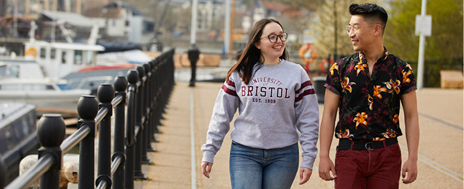 Two students walking along Bristol's harborside smiling at each other.