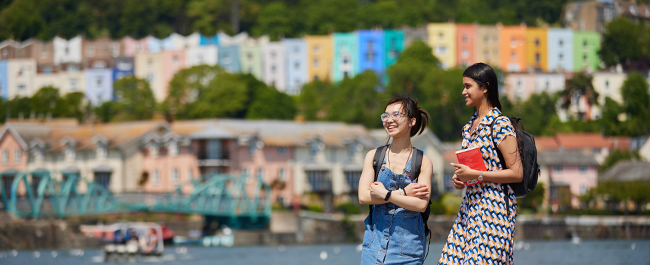 Two students standing on the harbourside smiling, the water, a foot bridge and a row of colourful houses in the background.