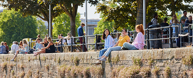 Three students sitting on a wall by the harbourside. Other people also sitting and enjoying the sunshine.