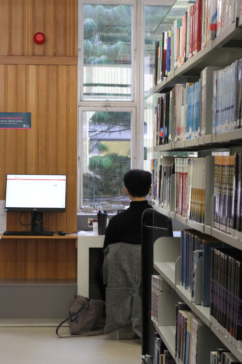 At the end of a book case is a student working on a desk which faces a window.