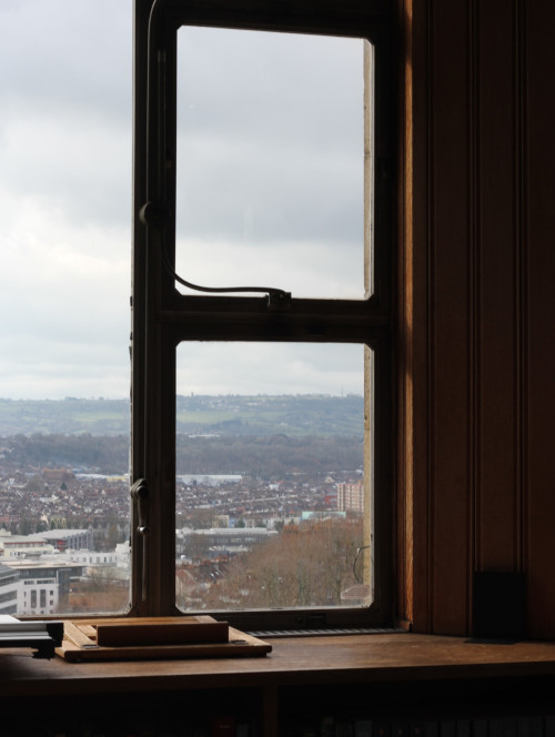 A small pile of books in front of a window frame, through the window in a view of Bristol and the skyline. 