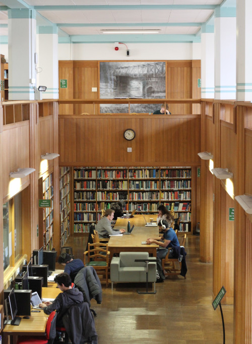 Looking down onto the ground floor of the Queen's library from the balcony, can see students working at computers and a group of students working at a wooden table. 