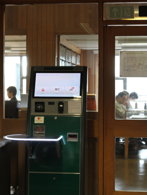 A machine used to take out books, behind it are glass panels which you can see through into the library where students are studying at desks. 
